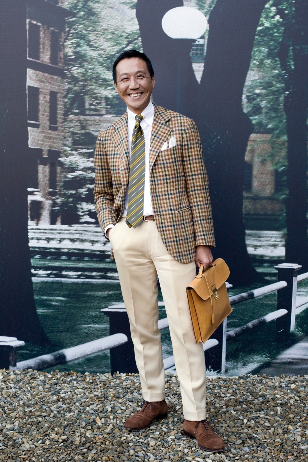 Yasuto Kamoshita of United Arrows wearing a tan checked blazer, cream trousers, striped tie and holding a tan leather briefcase.