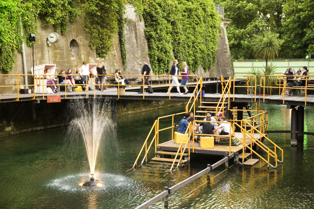 People gather on yellow-railed platforms and bridges beside a canal with a fountain, ivy-covered stone buildings in the background.