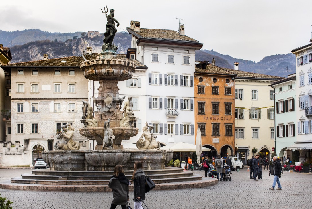 Neptune fountain statue in Piazza Duomo surrounded by historic buildings and Alpine mountains in Trento.