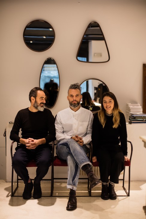 Three people sitting on chairs against a white wall with decorative mirrors.