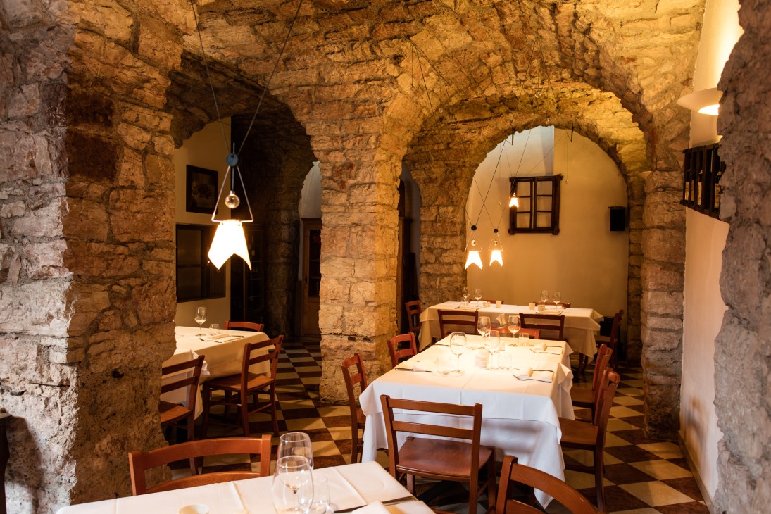Interior of Osteria Il Cappello showing stone arches, wooden chairs, white tablecloths, and pendant lighting.