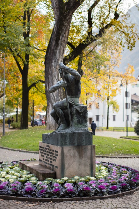 Bronze statue of a seated figure in Largo Luigi Pigarelli surrounded by colorful flowers and autumn foliage.