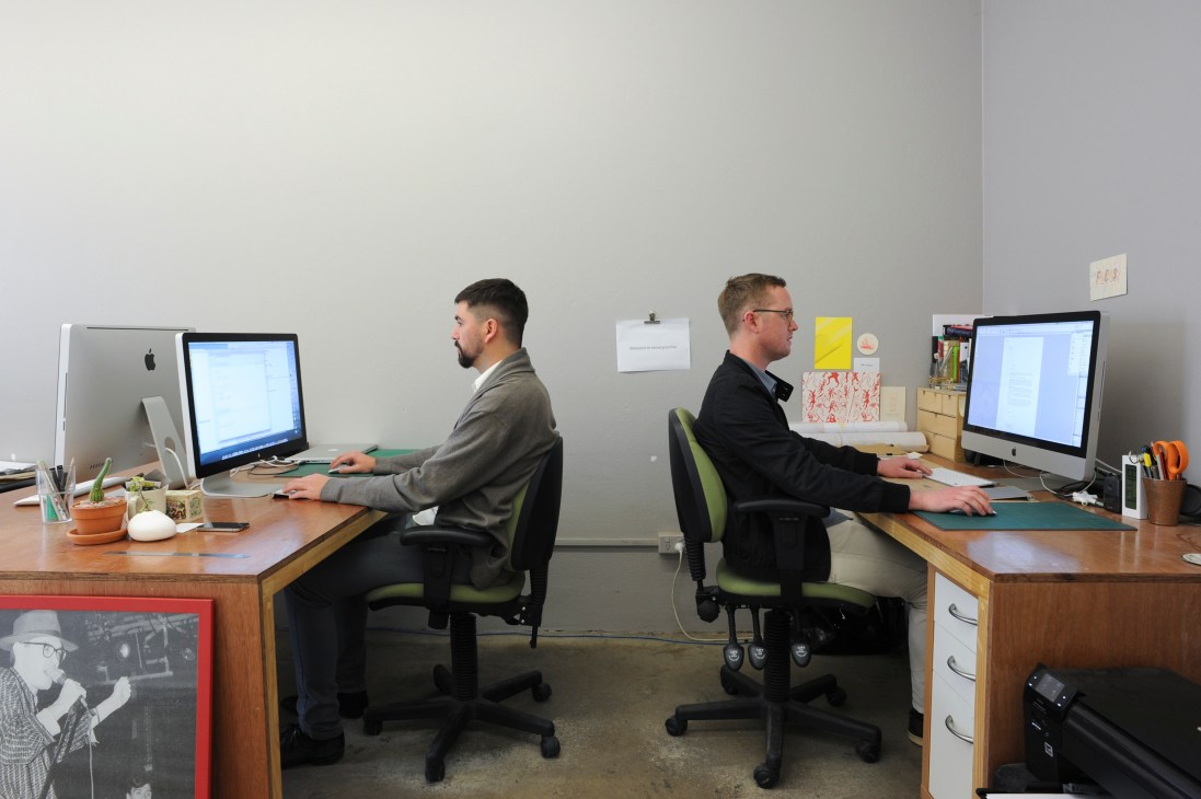 Two designers work at wooden desks in a minimalist office space with computers and potted plants.