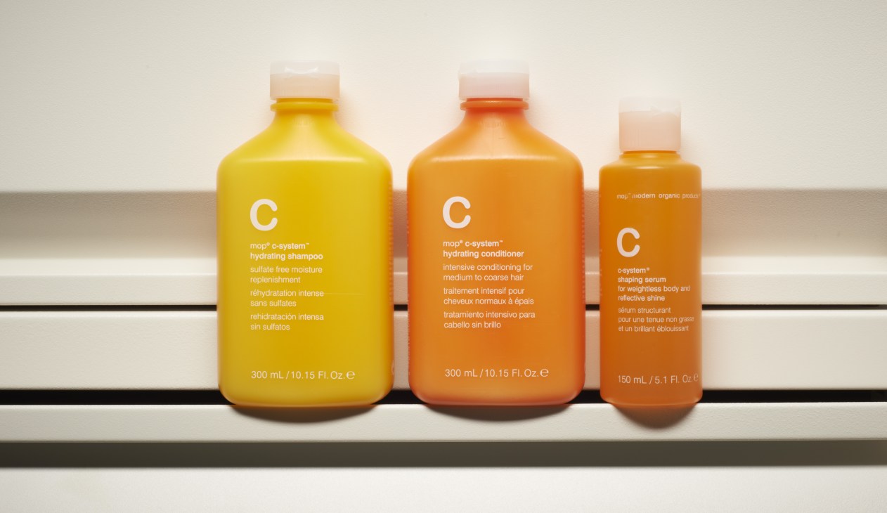 Three Mop* c-system hair products displayed: yellow hydrating shampoo, orange hydrating conditioner, and orange shaping serum bottles with white caps.