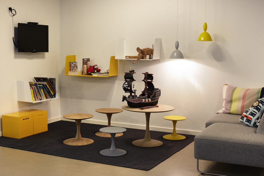 Children's corner with yellow storage boxes, wooden tables, toys, and colorful pendant lights designed by Muuto.