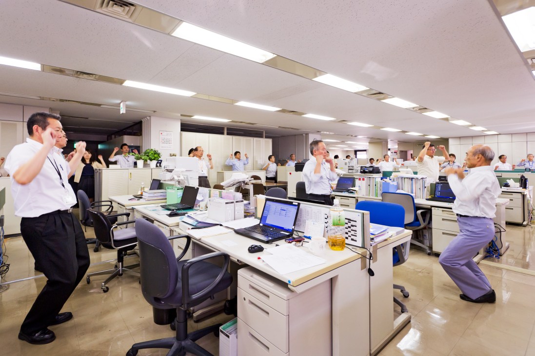 Office workers in business attire performing synchronized radio exercises at their desks in a modern workplace.