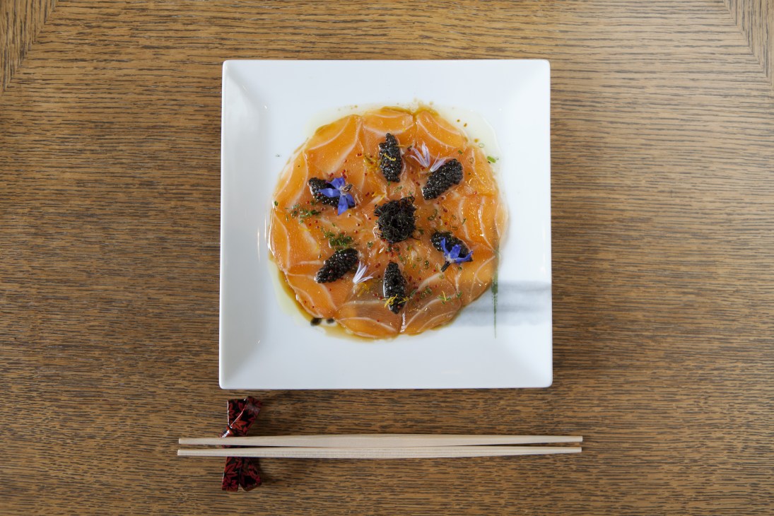 Salmon yuzu carpaccio plated on white square dish with caviar, lobster roe, herbs and edible flowers.