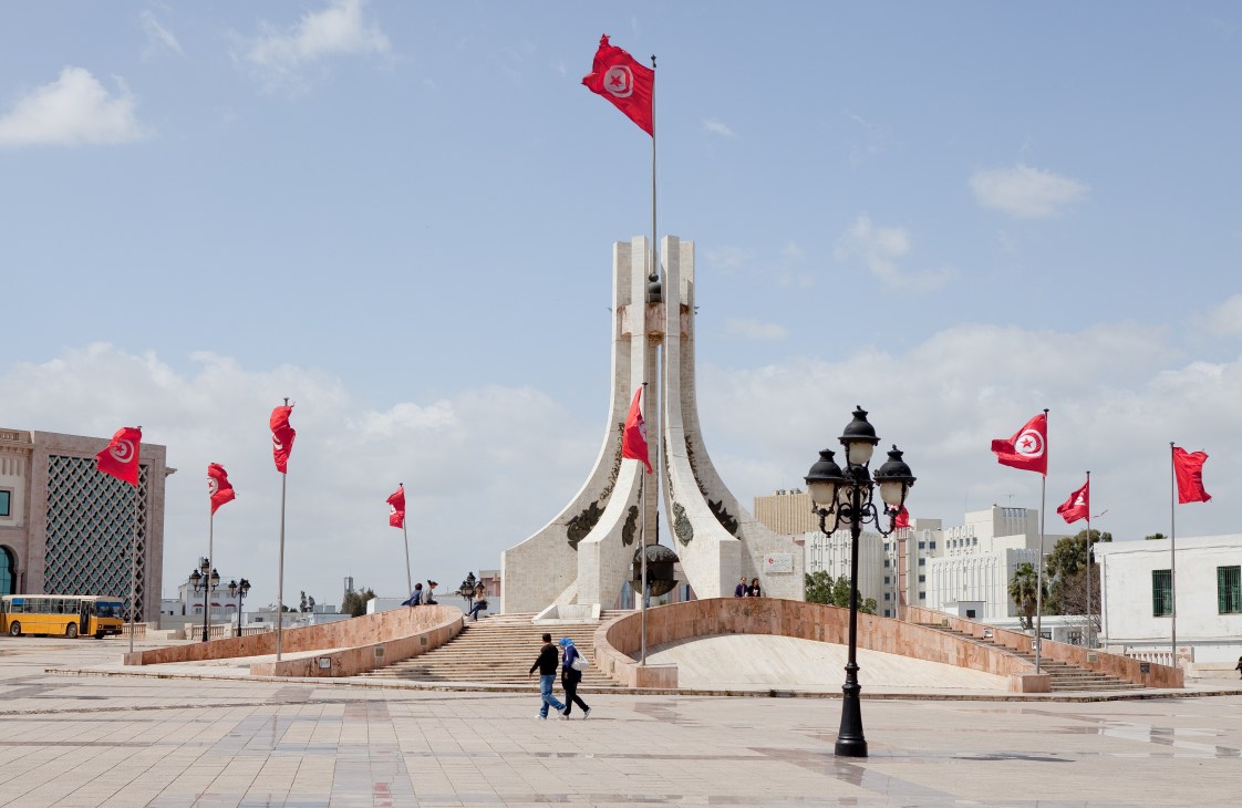 Tunisian flags fly around a modernist monument in a large public square with buildings in the background.