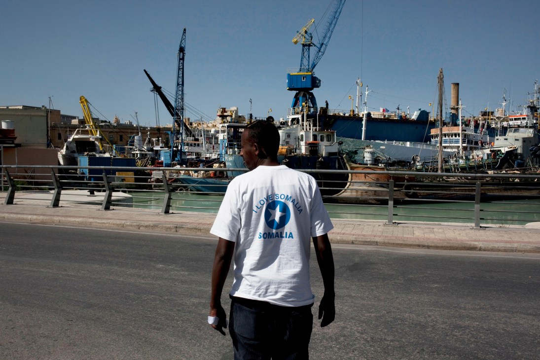 Man wearing a white "I Love Somalia" t-shirt standing at a harbor with cargo ships and cranes.