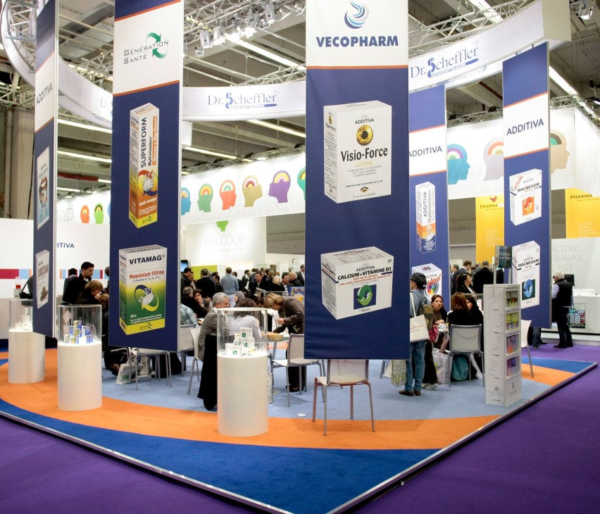 Pharmaceutical trade fair exhibition floor with branded booths displaying products and visitors.