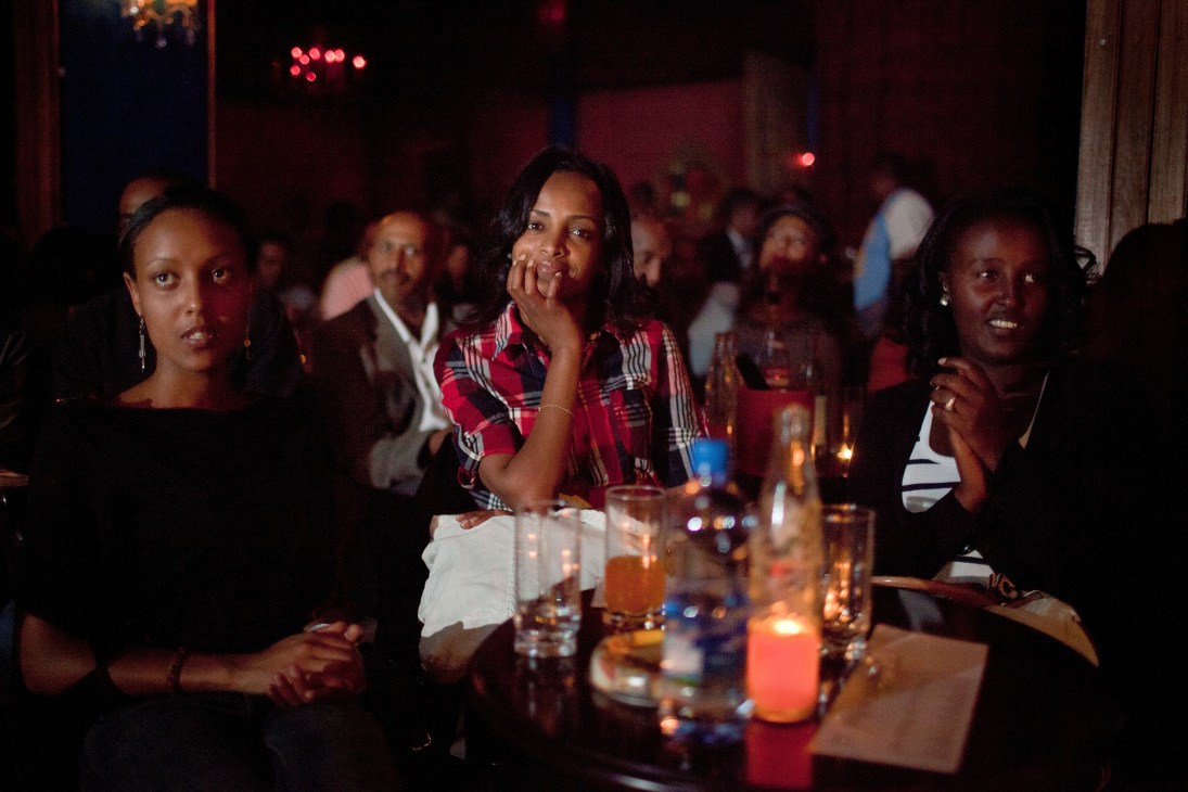 Audience members sitting at tables in a dimly lit jazz club with red ambient lighting and drinks.