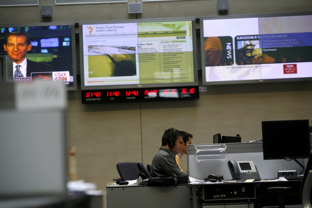 RFE/RL journalist wearing headphones at desk with multiple broadcast monitors displaying news content.