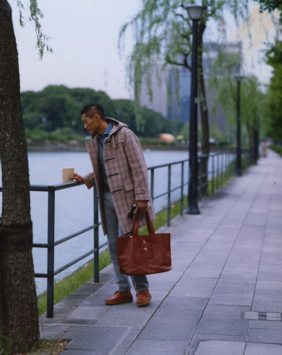 Man in plaid duffle coat and brown tote bag standing by a waterside railing in Tokyo.