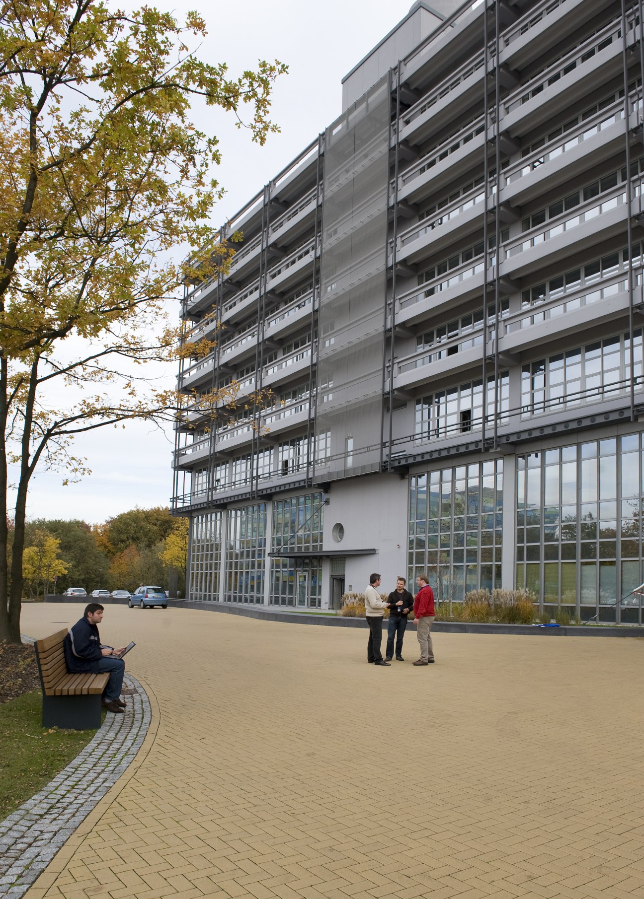 Modern multi-storey office building with glass and metal façade surrounded by autumn trees and paved plaza with people.