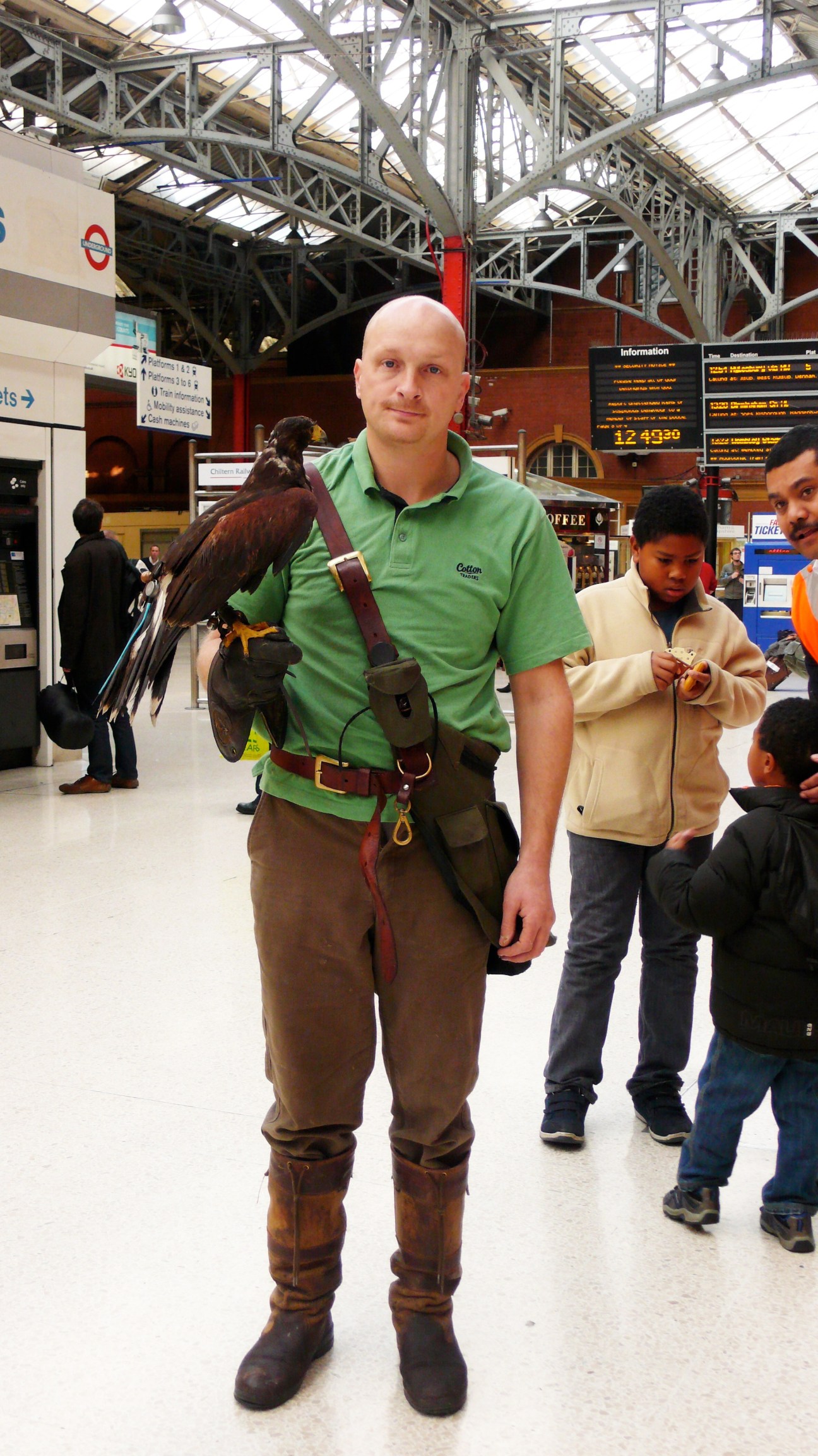 A Hawk Force falconer in a green polo shirt holds a trained hawk at a London train station.