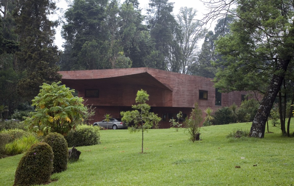 Modern red-brick building with curved roofline set in landscaped grounds with tall trees and manicured lawns.