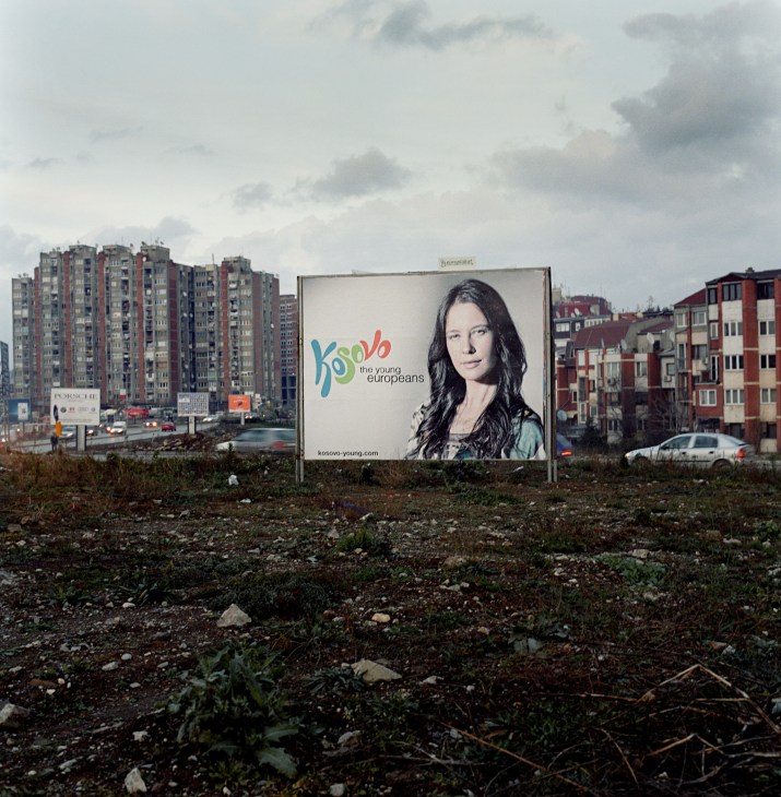 Kosovo's 'The Young Europeans' campaign billboard featuring a woman's portrait in a residential area with apartment buildings.