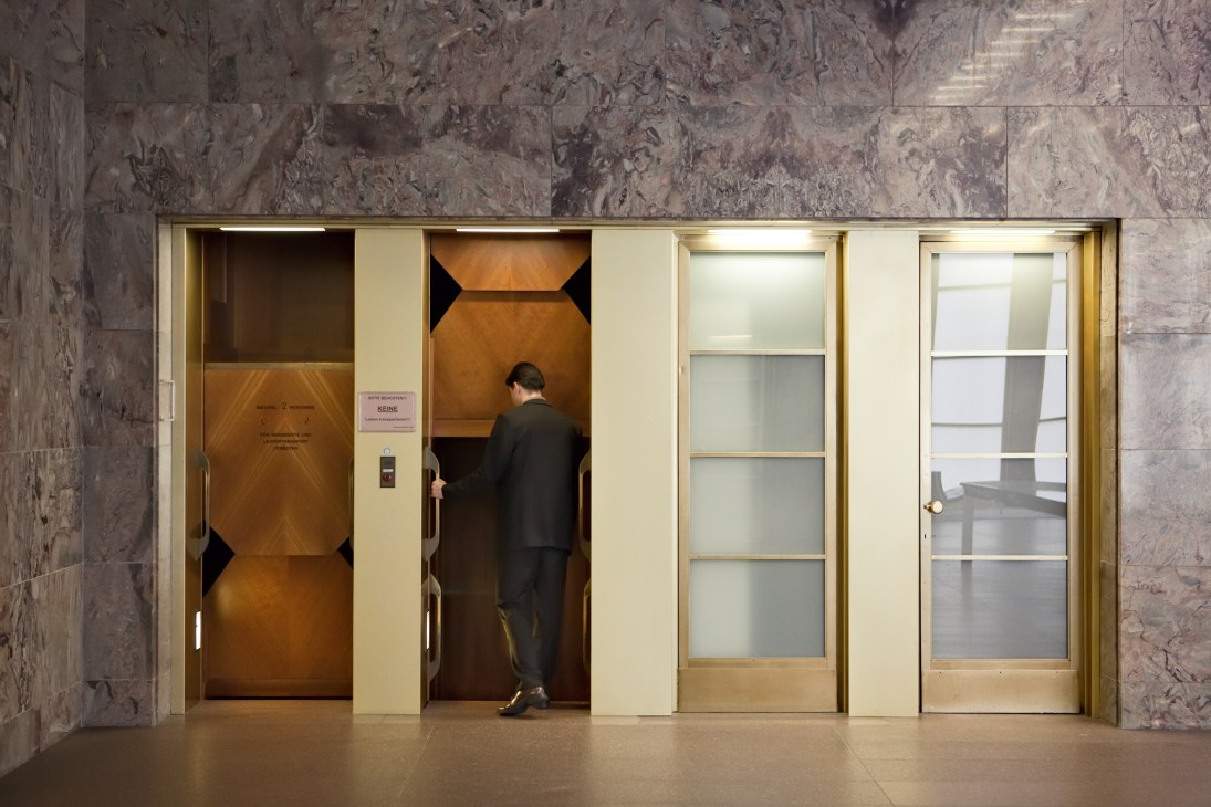Man in dark suit entering a vintage wooden paternoster lift in a marble-walled hall.