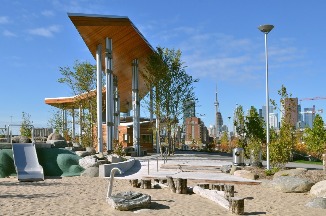 Modern wooden pavilion with green seating and wooden logs on a sandy beach with Toronto skyline in background.