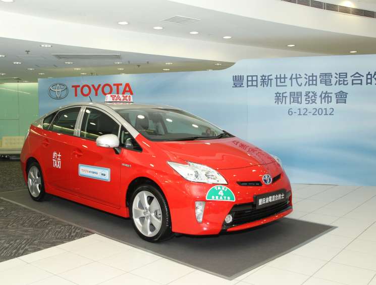 Red Toyota Prius hybrid taxi on display at a press event in Hong Kong with Chinese signage and Toyota branding visible.