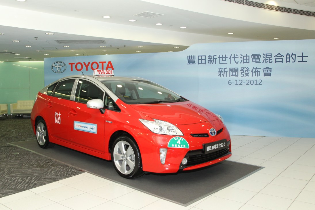 Red Toyota Prius hybrid taxi on display at a press event in Hong Kong with Chinese signage and Toyota branding visible.