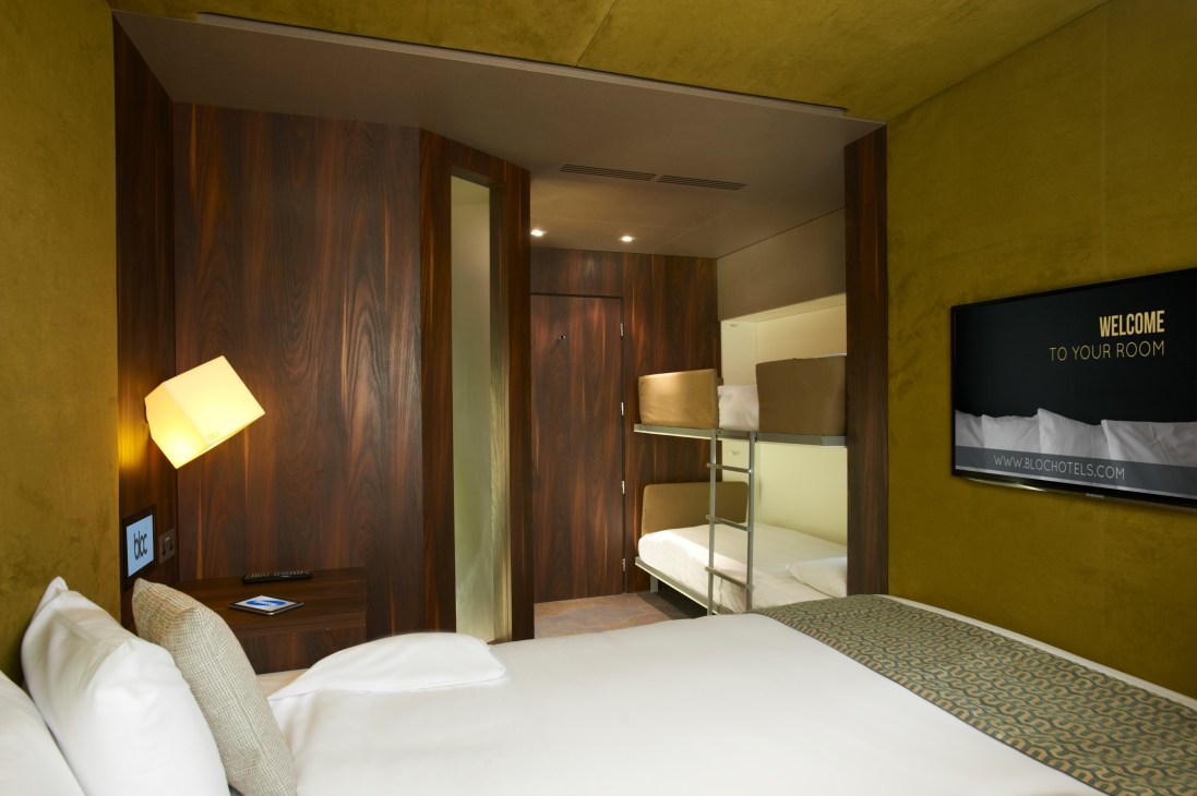 Modern budget hotel room with yellow walls, wood paneling, bed, and bunk beds with welcome sign.