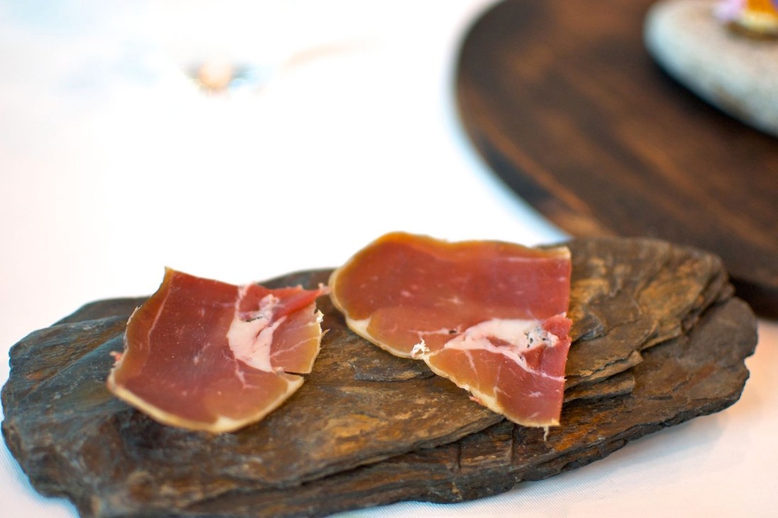 Sliced cured meat on a dark wooden board with tableware blurred in background.
