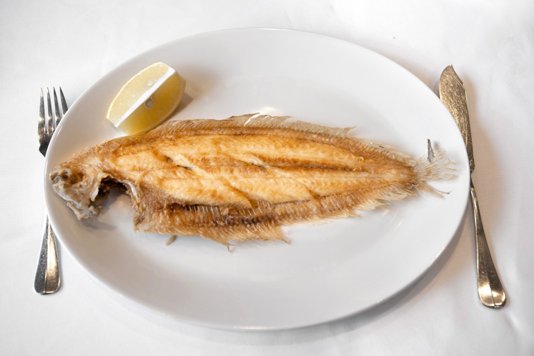 A whole Dover sole fried until golden, served on a white plate with lemon and cutlery.