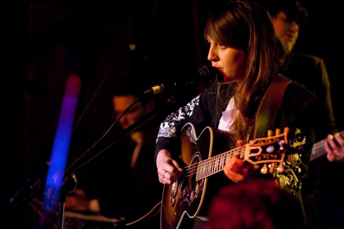Caitlin Rose performing with acoustic guitar at microphone on stage with colored stage lighting.