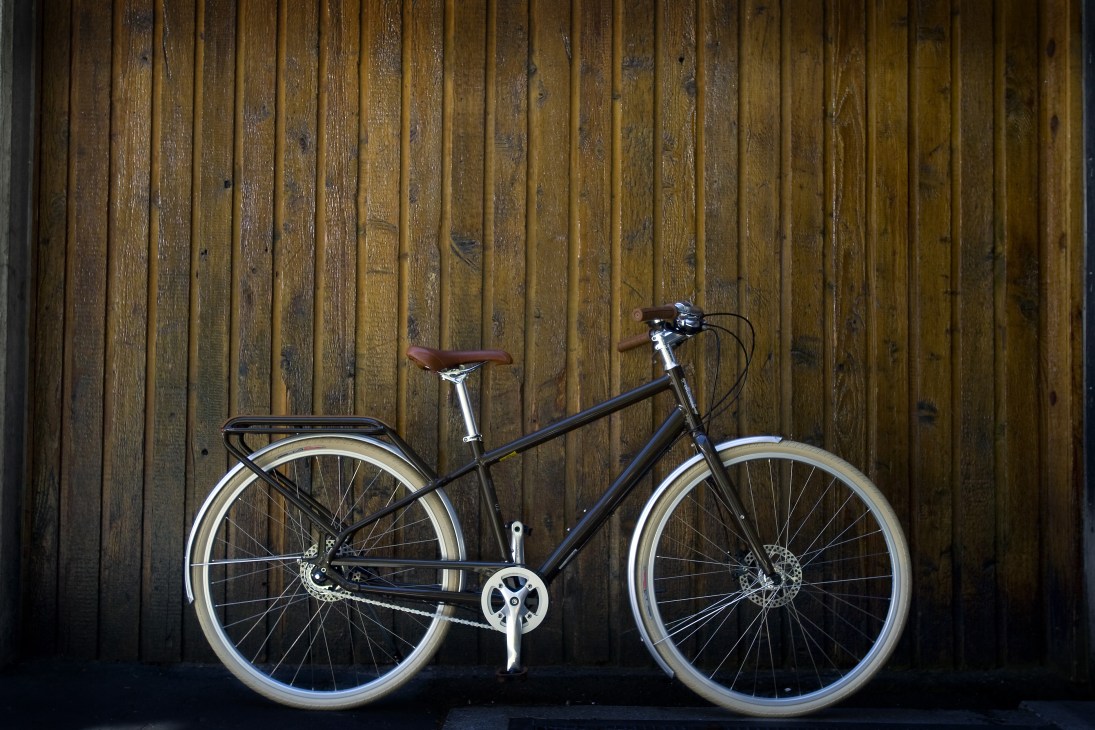 Black Globe HAUL commuter bike with brown saddle and cream-colored wheels against wooden wall.