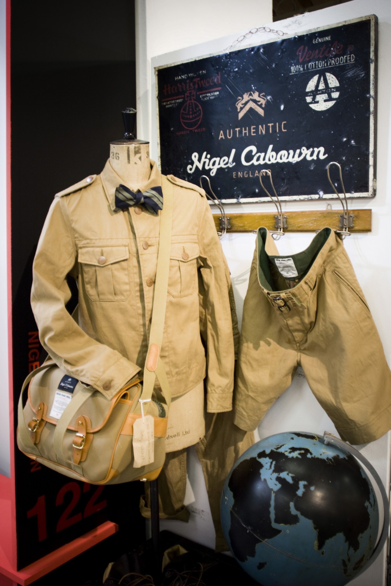 Mannequin displaying Nigel Cabourn's khaki military-inspired jacket with bow tie, shorts, and canvas bag against vintage signage.