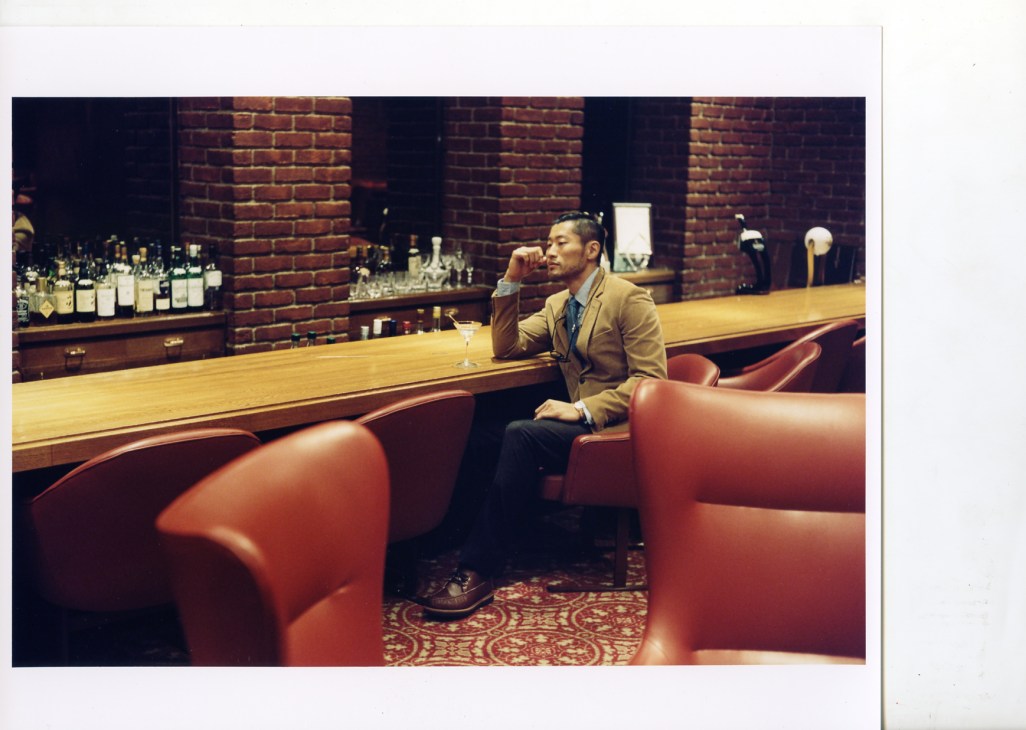 Man in Hackett jacket and tie sitting at bar counter with cocktail in 1970s lounge setting.