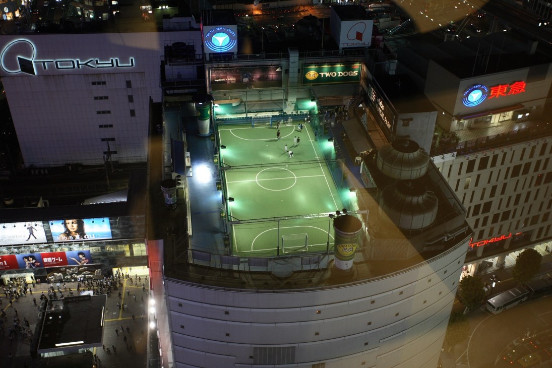 Aerial night view of Adidas Futsal Park, an illuminated rooftop soccer pitch on top of a Tokyo department store surrounded by city buildings.