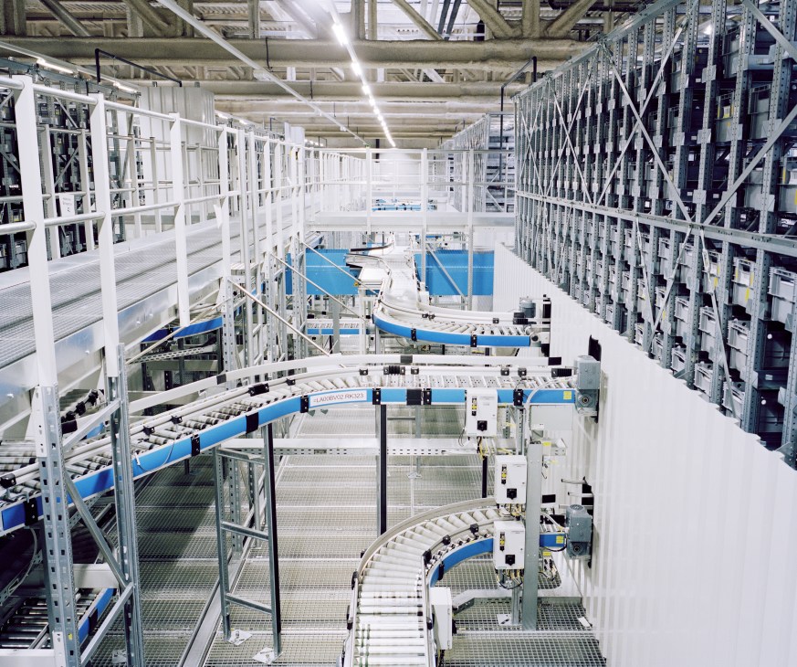 Fully automated high-bay racking system with white metal shelving and blue conveyor belts in a Swiss Post sorting facility.