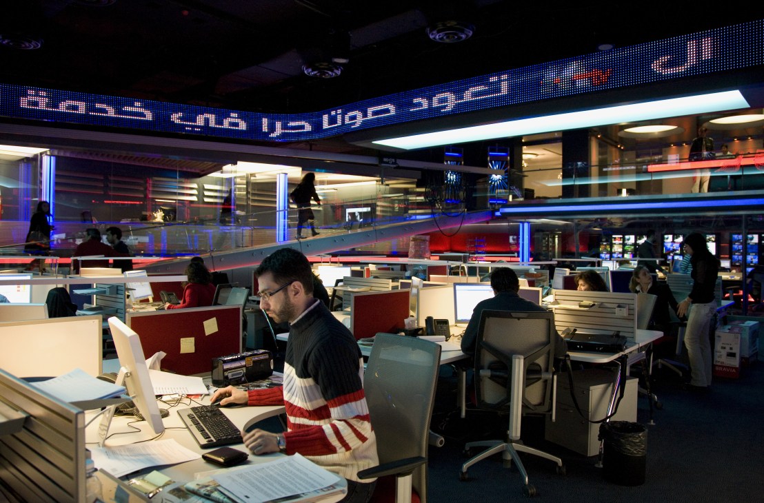 M TV newsroom with journalists working at desks beneath illuminated Arabic text signage and blue-red studio lighting.