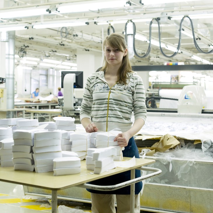 Woman in striped cardigan stands at workstation with stacked white underwear packages in Milavitsa factory.