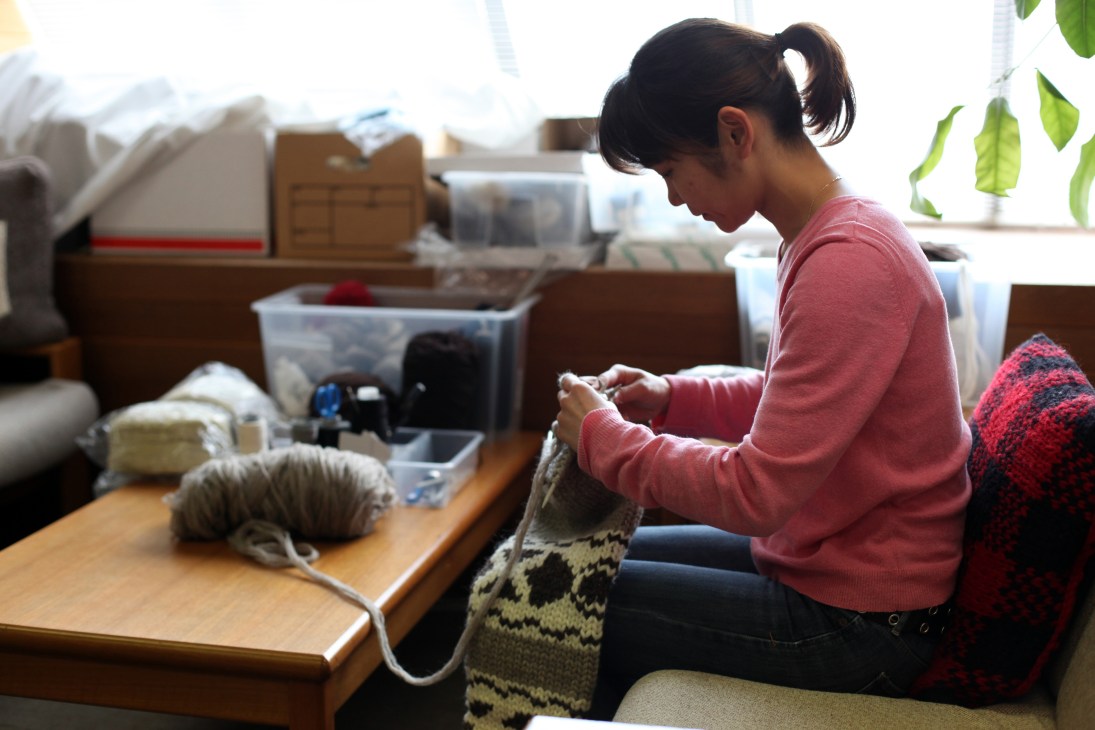 Woman in pink sweater hand-knitting a patterned sweater at a wooden table in a bright studio.