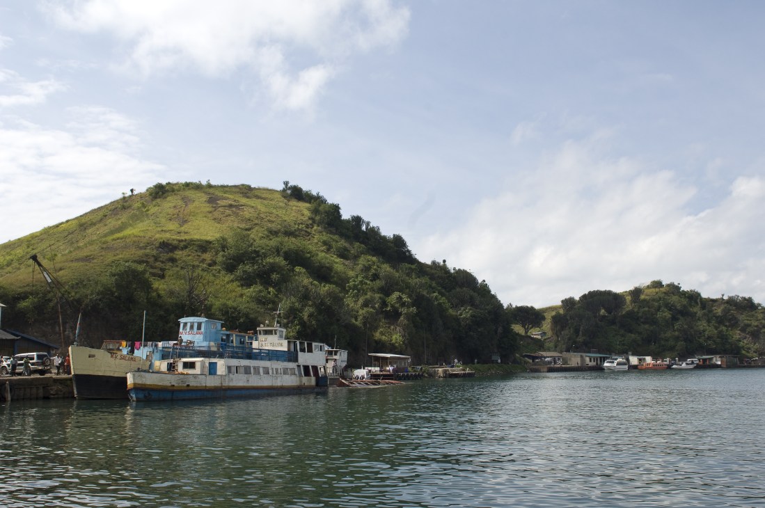 Blue and white cargo vessel moored at Kivu Harbour with lush green hills and vegetation in the background.