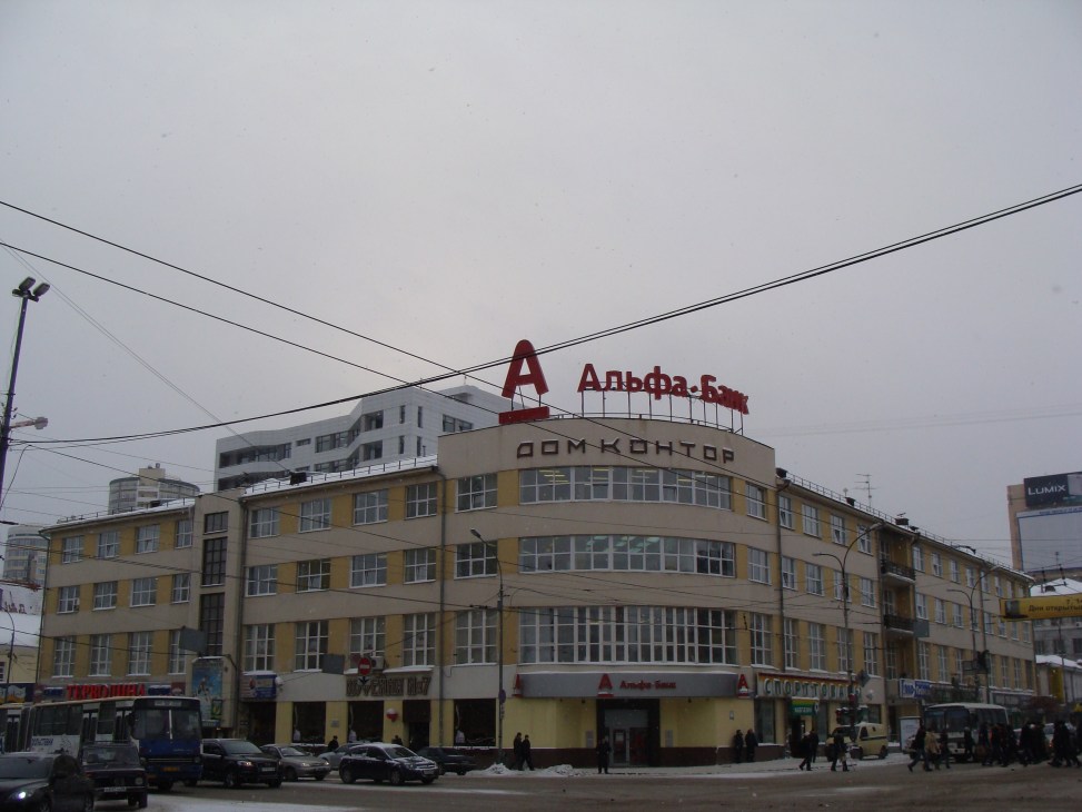 Yellow and cream-colored Constructivist building with Alfa Bank signage and modern office tower behind it in snowy Ekaterinburg.
