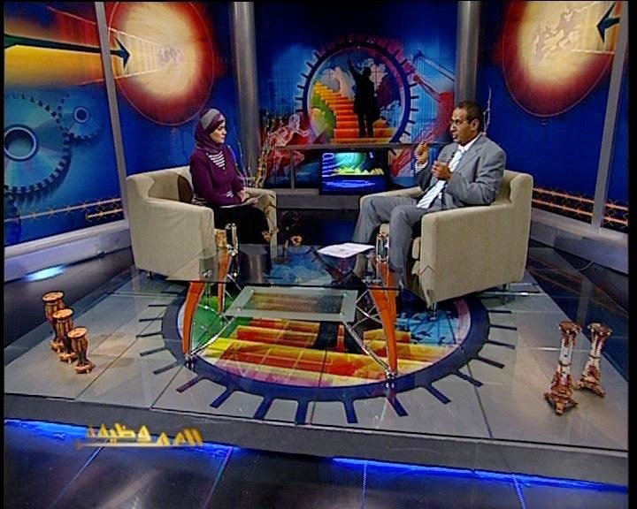Two people sitting in chairs on a colorful studio set with futuristic backdrop and lighting.