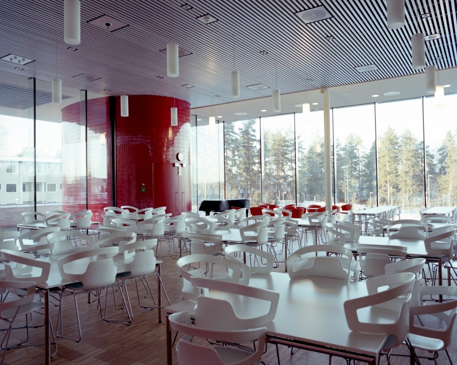Modern hospital cafeteria with white Alias chairs, red cylindrical structure, and floor-to-ceiling windows overlooking forest.