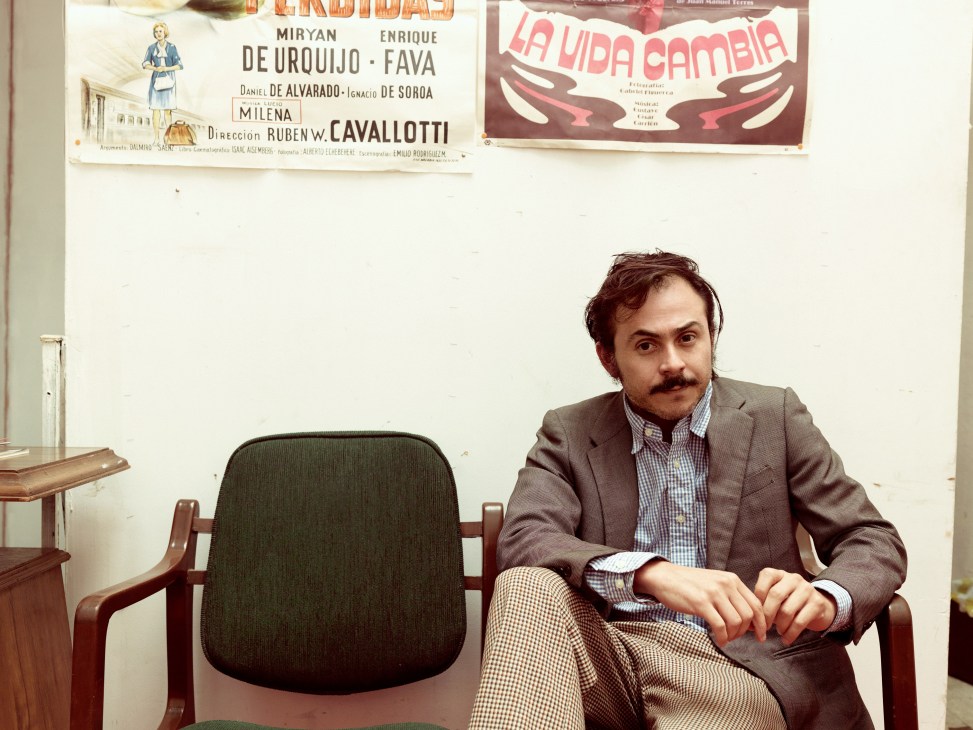 Director Gerardo Naranjo sitting in an office chair with vintage Mexican film posters on the wall behind him.