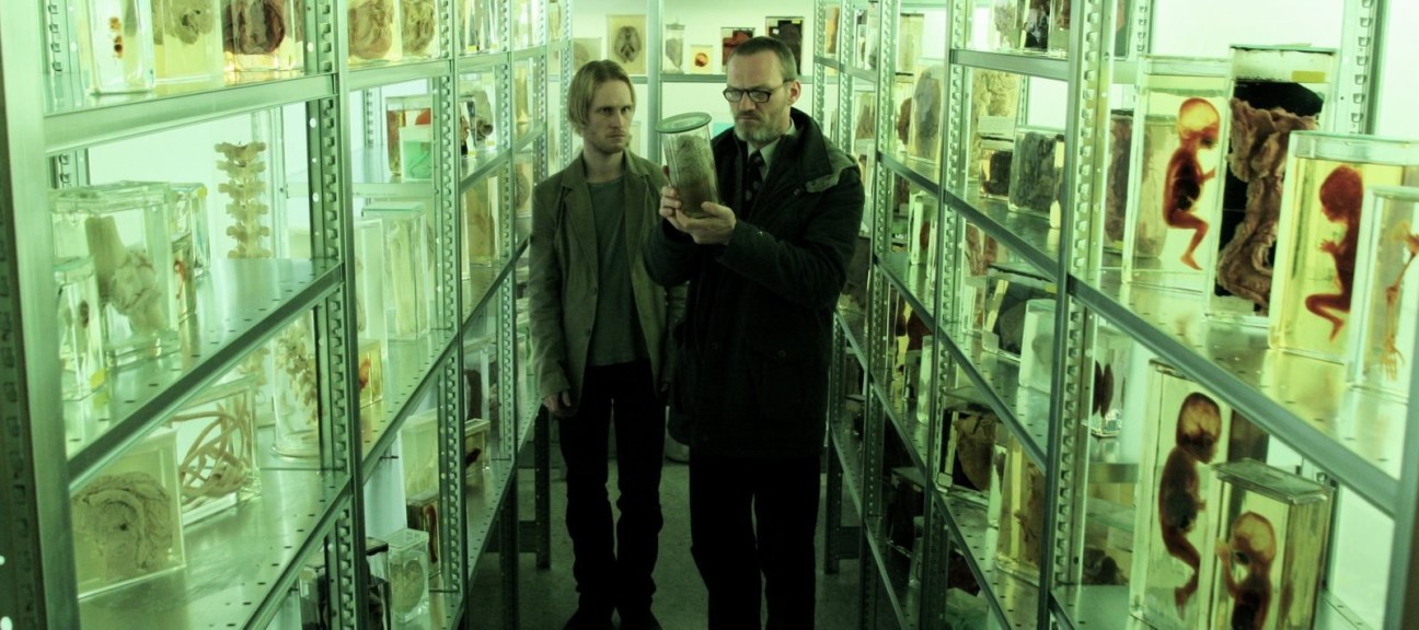 Two men walk through a museum corridor lined with glass display cases containing specimens under green lighting.