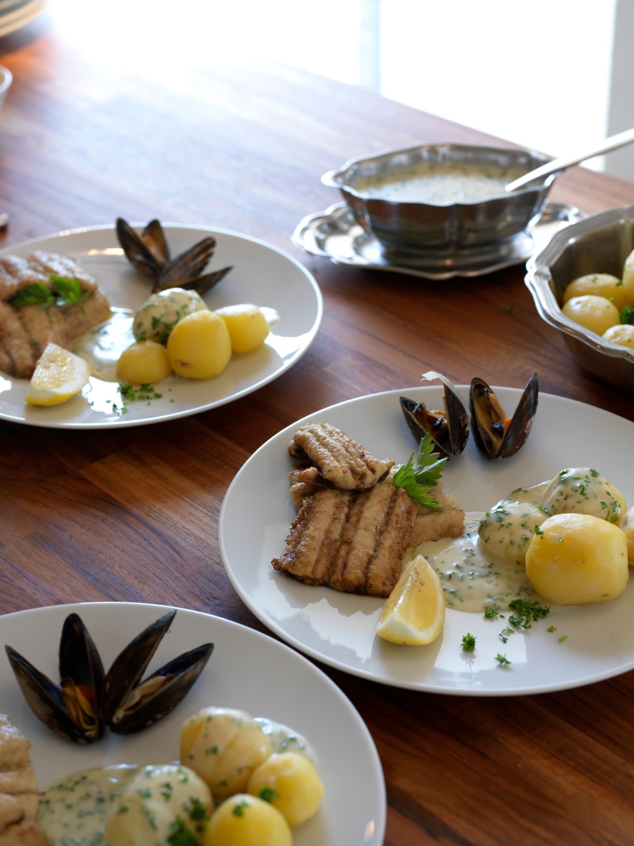 Plated pan-fried garfish with boiled potatoes, parsley gravy, lemon wedges and mussels on white plates.