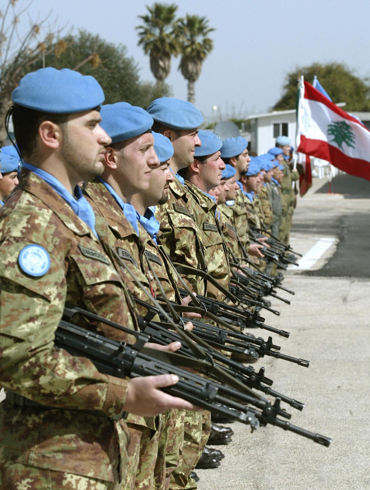 UN Italian peacekeeping troops in blue berets stand in formation holding rifles with Lebanese flag in background.