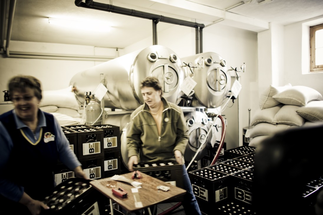 Two women prepare beer bottles for shipping at Bieraria Tschlin brewery with fermentation vats in background.