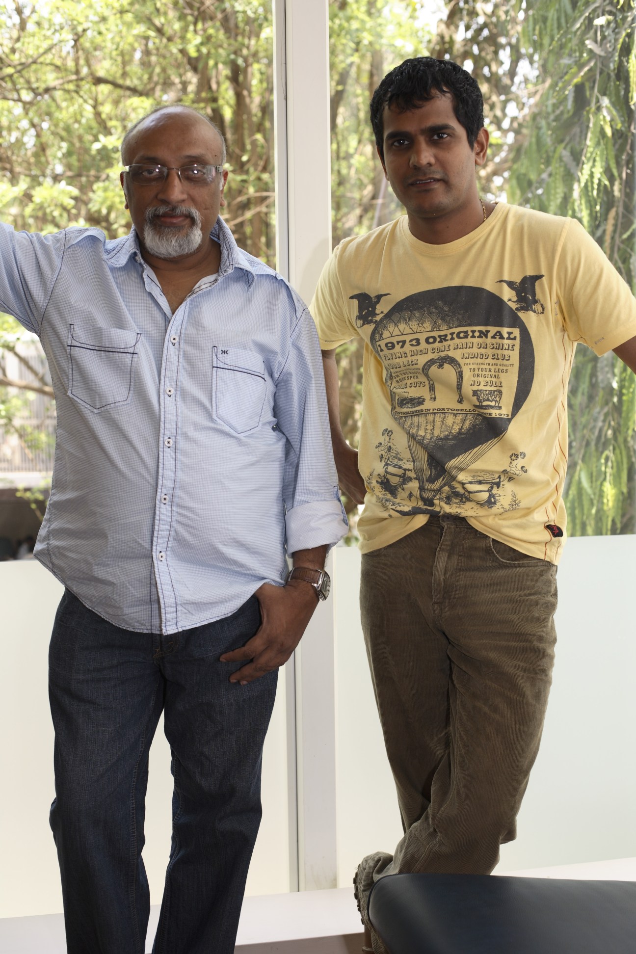 Two men stand together in an office with windows and greenery behind them; one wears a light blue shirt and glasses, the other wears a yellow graphic t-shirt.