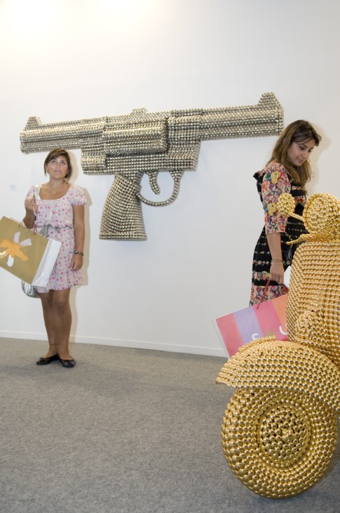 Two women stand near a revolver and coiled snake sculpture made of metallic beads by Valay Shende.