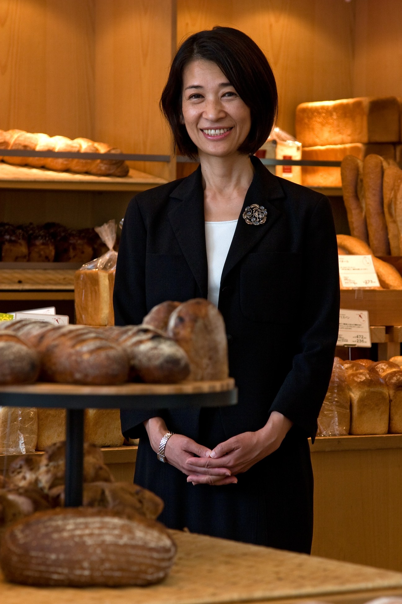 Masako Yoshida smiles in an Andersen bakery surrounded by fresh bread and pastries on wooden shelves.