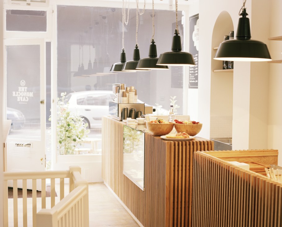 Monocle Café counter with red oak panelling, black pendant lights, and display of coffee cups and food items.